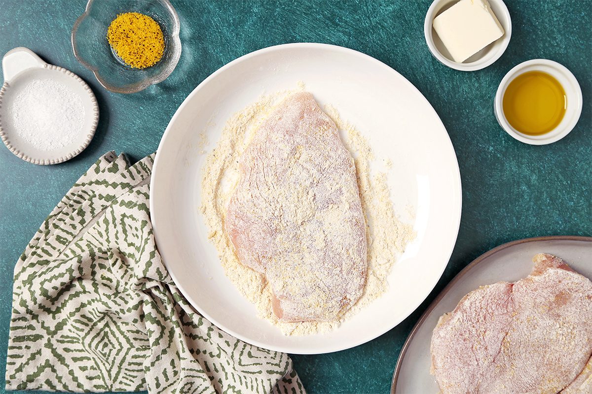 A raw chicken breast coated in flour sits on a white plate. Nearby are ingredients: a bowl of seasoning, a dish of salt, a small bowl of butter, a container of olive oil, and a patterned cloth on a green surface.