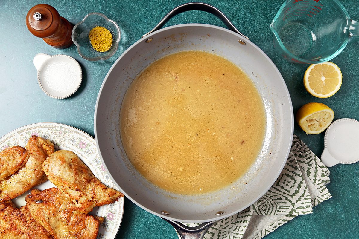A large pan filled with brown gravy is on a green surface next to a patterned cloth. To the left, fried chicken pieces are on a plate. Surrounding the pan are a pepper mill, small bowls with spices, a lemon half, a glass pitcher, and a salt container.