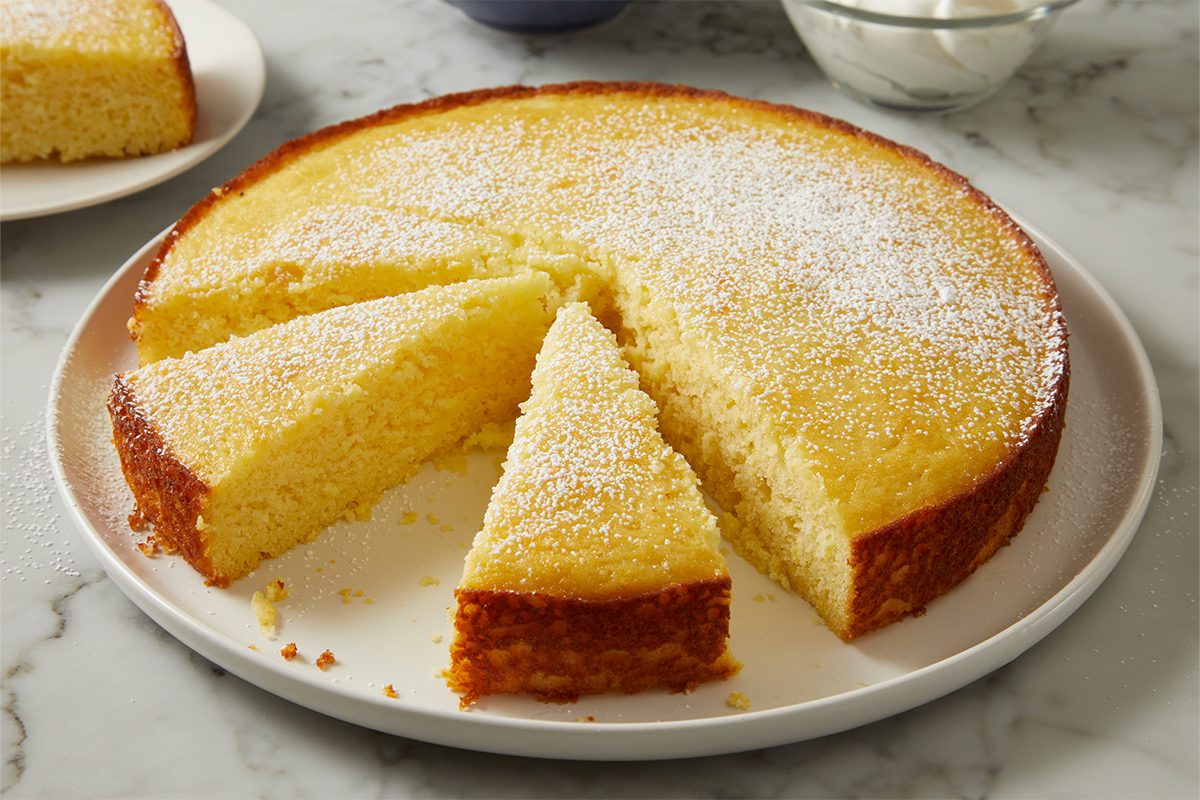 Side view of baked lemon ricotta cake on a serving plate with cut slices dusted with confectioners' sugar on a white marble countertop.