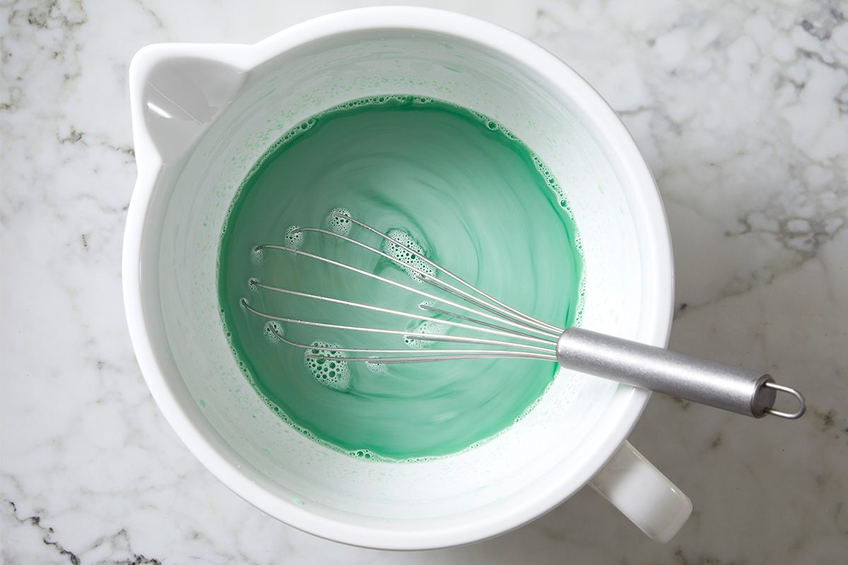 Overhead view of a large white bowl with a whisk with the lime gelatin mix and boiling water being mixed on a white marble countertop.