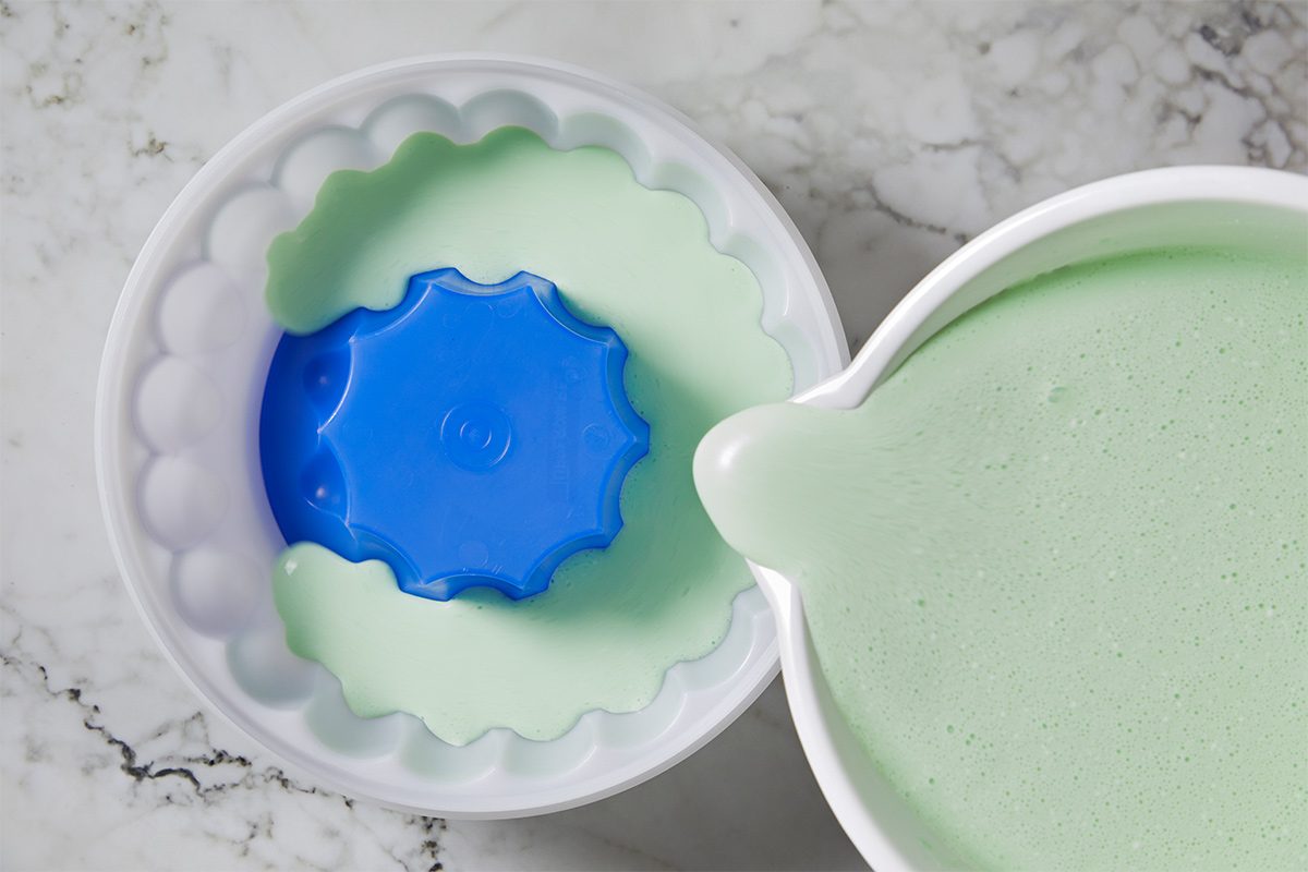 Overhead view of the lime sherbet, lime gelatin and whipped topping mixture in the large bowl being poured into a white 8-cup ring mold on a while marble countertop.
