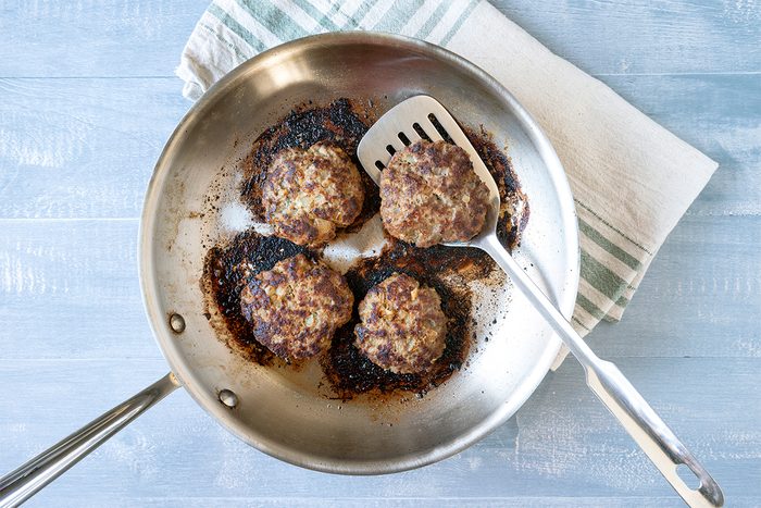 Four browned meat patties in a stainless steel frying pan with a spatula resting inside. The pan sits on a light blue wooden surface with a folded striped cloth underneath.