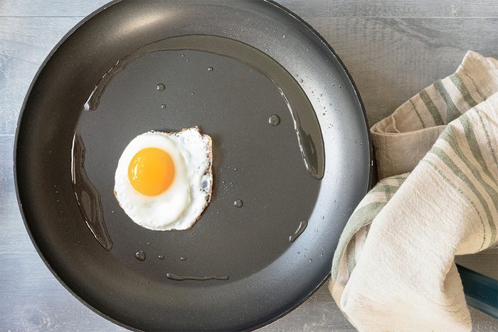 A single sunny-side-up egg is frying in a black non-stick pan on a gray surface. A striped cloth is placed nearby.