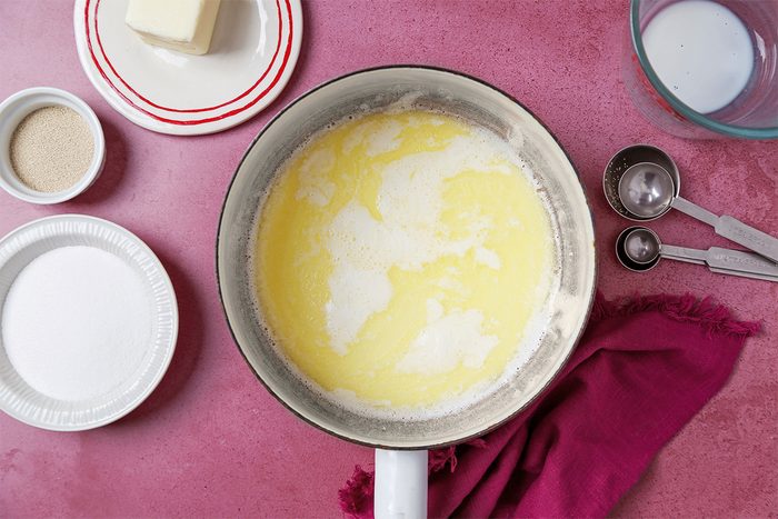 A mixing bowl with a creamy yellow batter on a pink surface, surrounded by ingredients like butter on a plate, a bowl of sugar, measuring cups, and a red cloth.