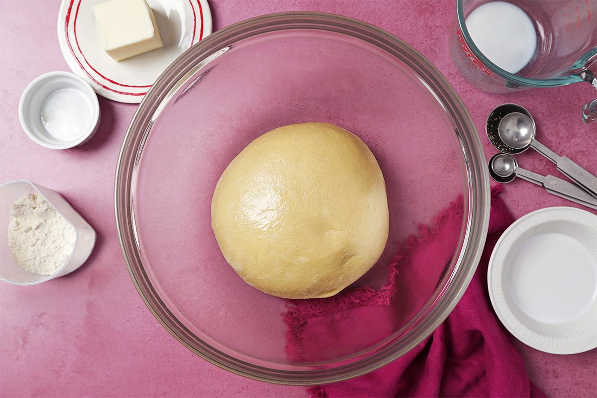 A ball of dough rests in a large glass bowl on a pink countertop, surrounded by ingredients like butter, flour, salt, measuring spoons, and a red cloth.