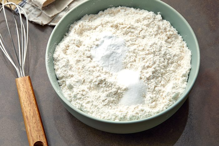 a close-up of a green bowl filled with white flour, two white piles of what looks like baking soda or salt, and a whisk with a brown wooden handle, the whisk is partially out of frame the bowl is sitting on a brown countertop