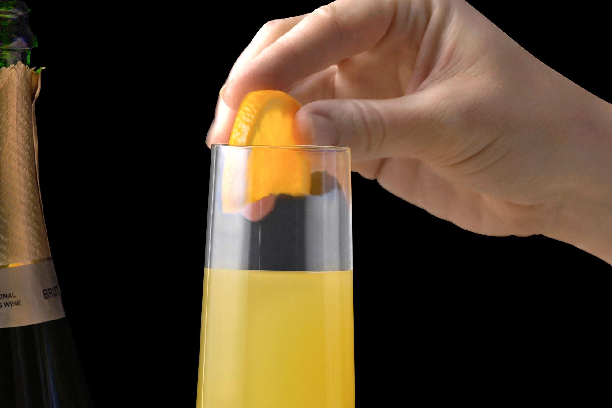 closeup shot of a hand gently placing a slice of orange into a glass of champagne; a bottle of sparkling wine is in the background to the left of the image; the background is black