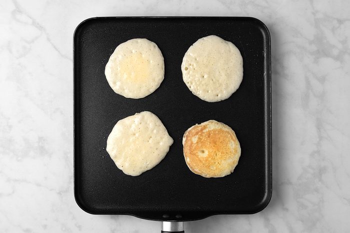 Four pancakes cooking on a black griddle, with three partially cooked and one fully cooked with a golden-brown color. The griddle is on a marble surface.