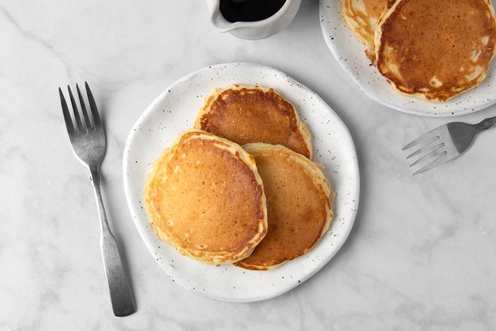 A white plate with three fluffy pancakes on a marble surface, accompanied by a fork. A jug of syrup and another plate with more pancakes are in the background.