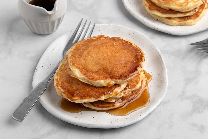 A stack of three golden-brown pancakes on a white plate, drizzled with syrup. A fork rests on the left side of the plate. In the background, there's a small pitcher with more syrup and another plate of pancakes.