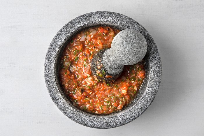 A gray stone mortar and pestle containing freshly made salsa with tomatoes, herbs, and spices. The mixture appears vibrant and slightly chunky against the smooth, speckled surface of the mortar.