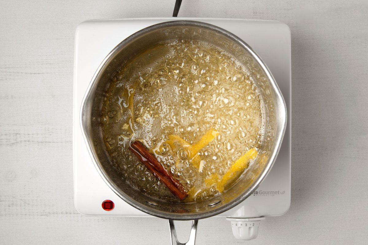 overhead shot of a pot of liquid simmering on a white stovetop; the pot is made of stainless steel and has a silver handle; the liquid inside is a pale golden color with visible bubbles, and there is a piece of cinnamon stick submerged in it;