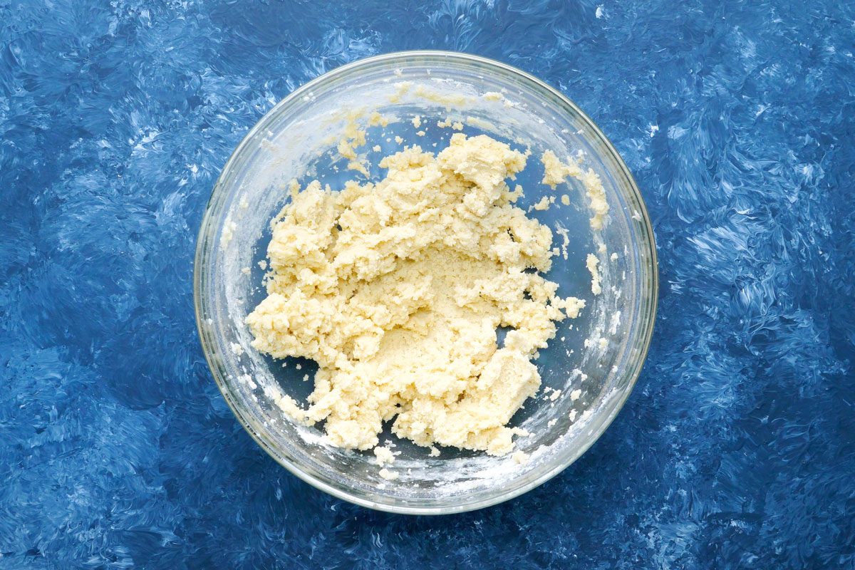 overhead shot of a clear glass bowl filled with a light yellow dough-like substance; the bowl sits on a blue surface with white streaks and swirls