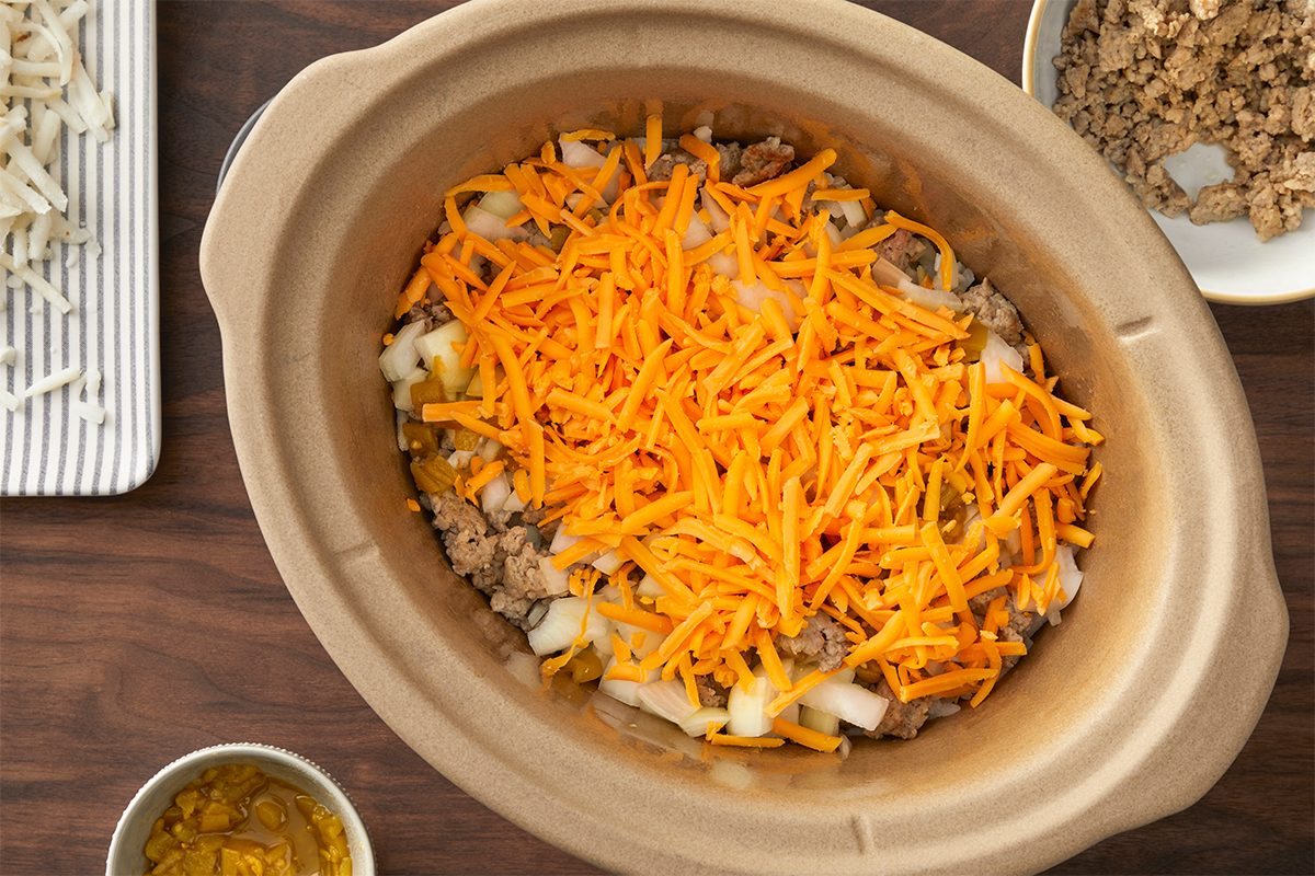 A slow cooker filled with shredded cheddar cheese layered over cooked ground meat and diced onions. To the side, there's a plate with more ingredients and a bowl with additional ground meat.