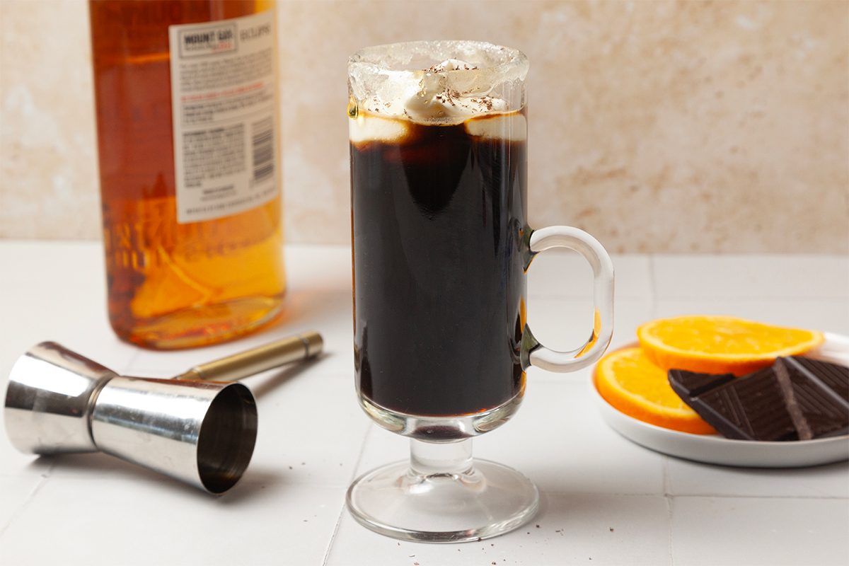 Glass mug filled with dark coffee topped with whipped cream, next to an orange slice and chocolate chunks on a plate. A liquor bottle and metal jigger are in the background on a light-colored surface.