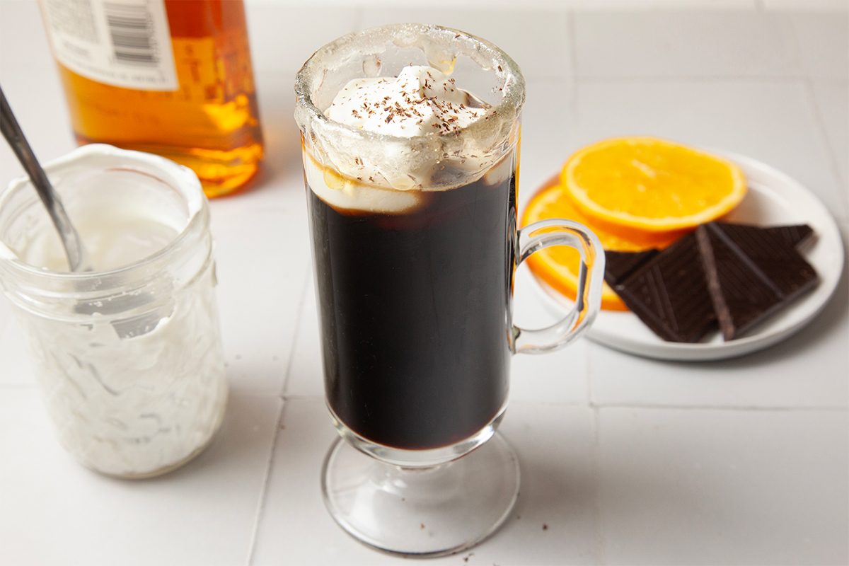 A glass of Irish coffee topped with whipped cream and chocolate shavings on a table. Nearby, there's a jar with a spoon, a bottle, and a plate with orange slices and dark chocolate squares.