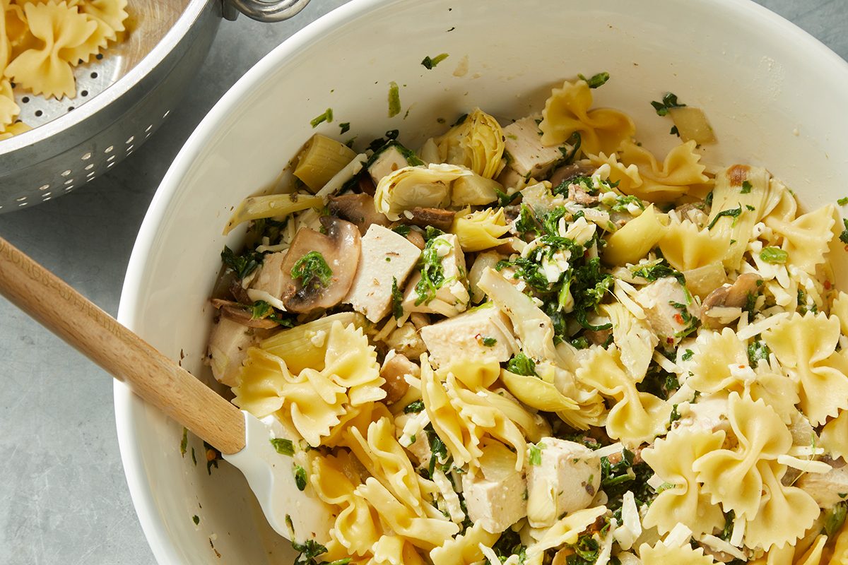 A white bowl filled with bowtie pasta mixed with chunks of chicken, mushrooms, artichoke hearts, and herbs. A wooden spoon rests in the bowl, and a metal colander is partially visible in the background on a gray surface.