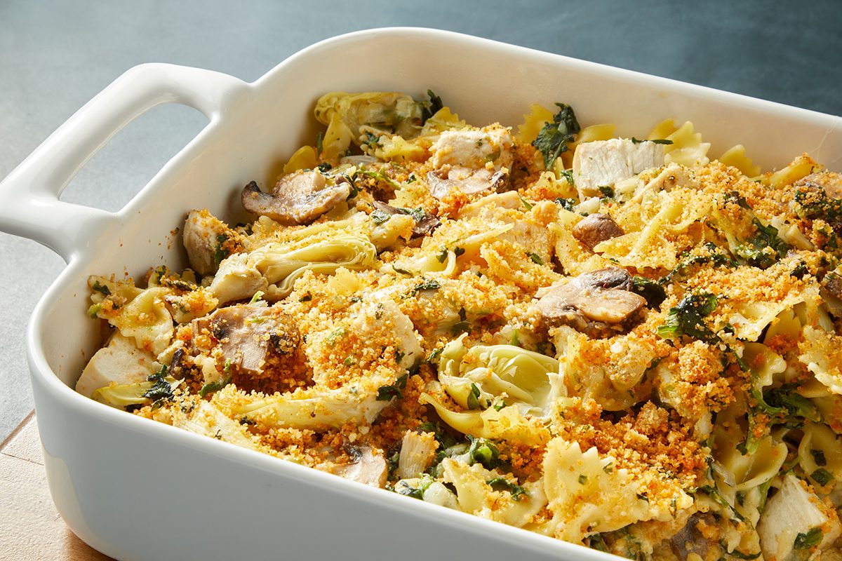 A white casserole dish filled with baked pasta, mushrooms, artichoke hearts, and leafy greens, topped with a crispy breadcrumb crust. The dish is set on a wooden surface.
