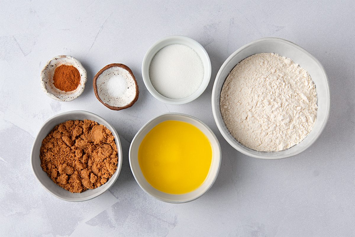 Bowls containing cocoa powder, melted butter, flour, milk, baking powder, and cinnamon are arranged on a light gray surface.