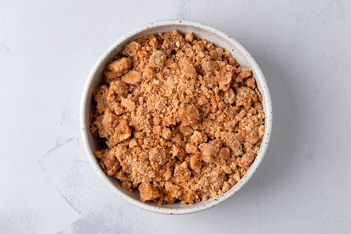 A bowl filled with crumbled brownish topping or mixture, possibly resembling streusel or a crumbly dessert component, placed on a light gray surface.