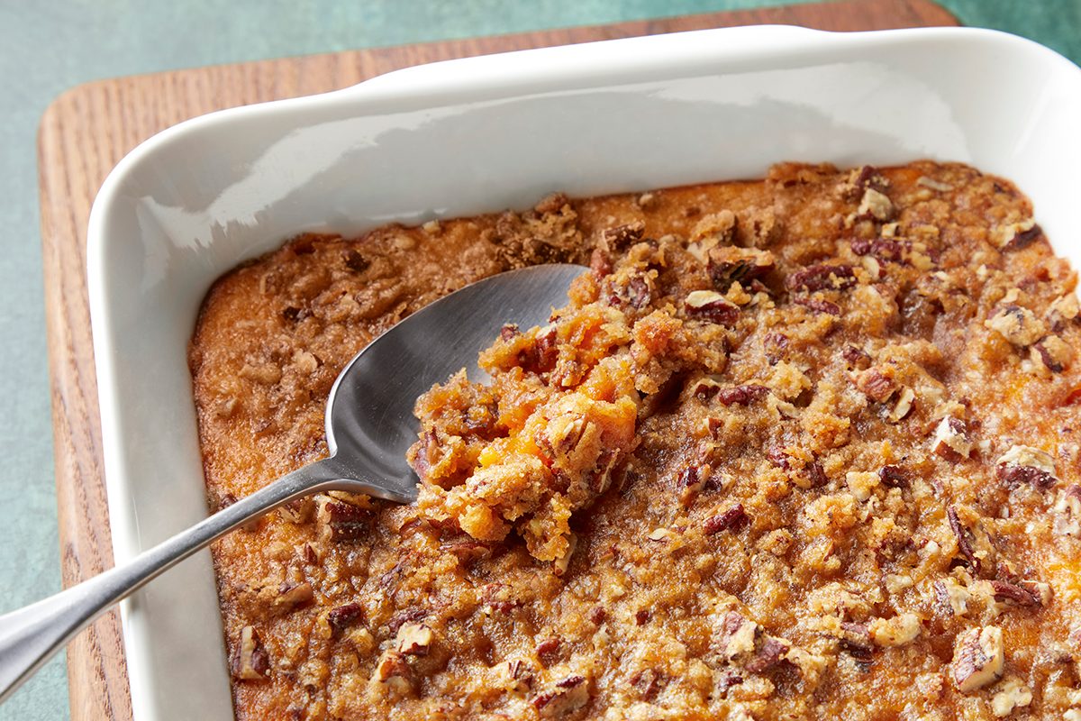 A white baking dish holds a sweet potato casserole topped with a crispy pecan streusel. A silver spoon is scooping a portion from the corner, revealing the creamy orange filling beneath the crust. The dish rests on a wooden surface.