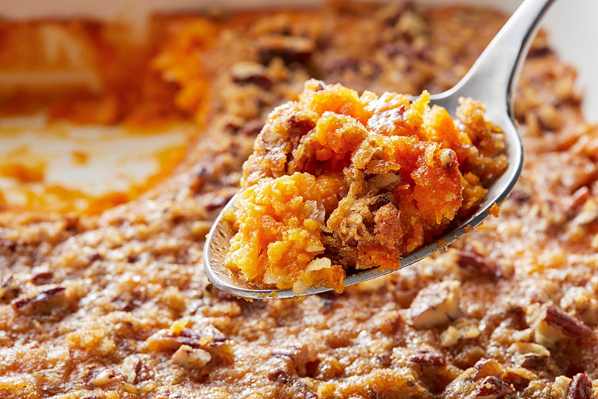 A close-up of a spoonful of sweet potato casserole topped with a crumbly pecan mixture. The background shows the dish with a portion already served, revealing the casserole's fluffy orange interior.
