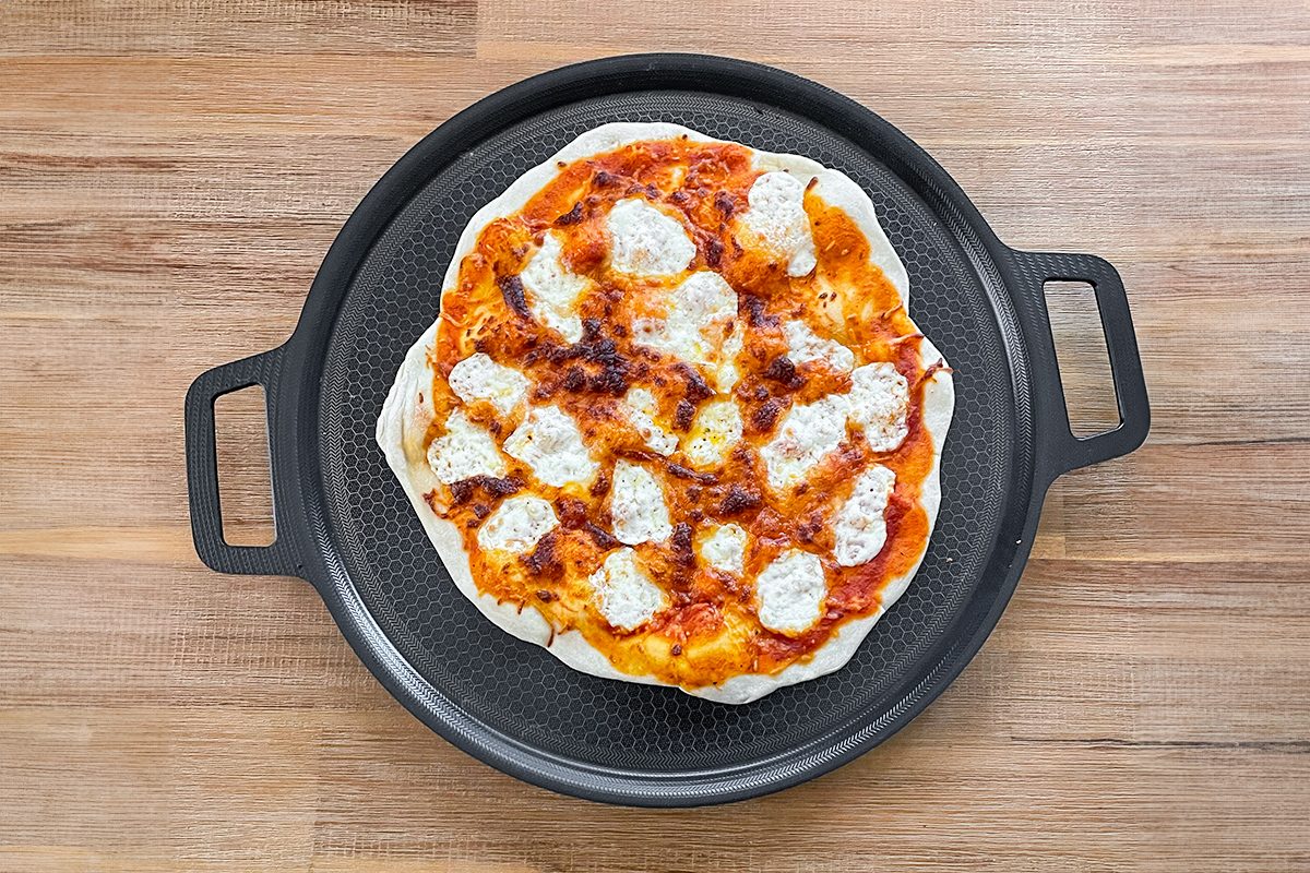 A homemade pizza with a golden crust, topped with melted cheese and tomato sauce, on a round pizza pan. The pan has two handles and is placed on a wooden table.