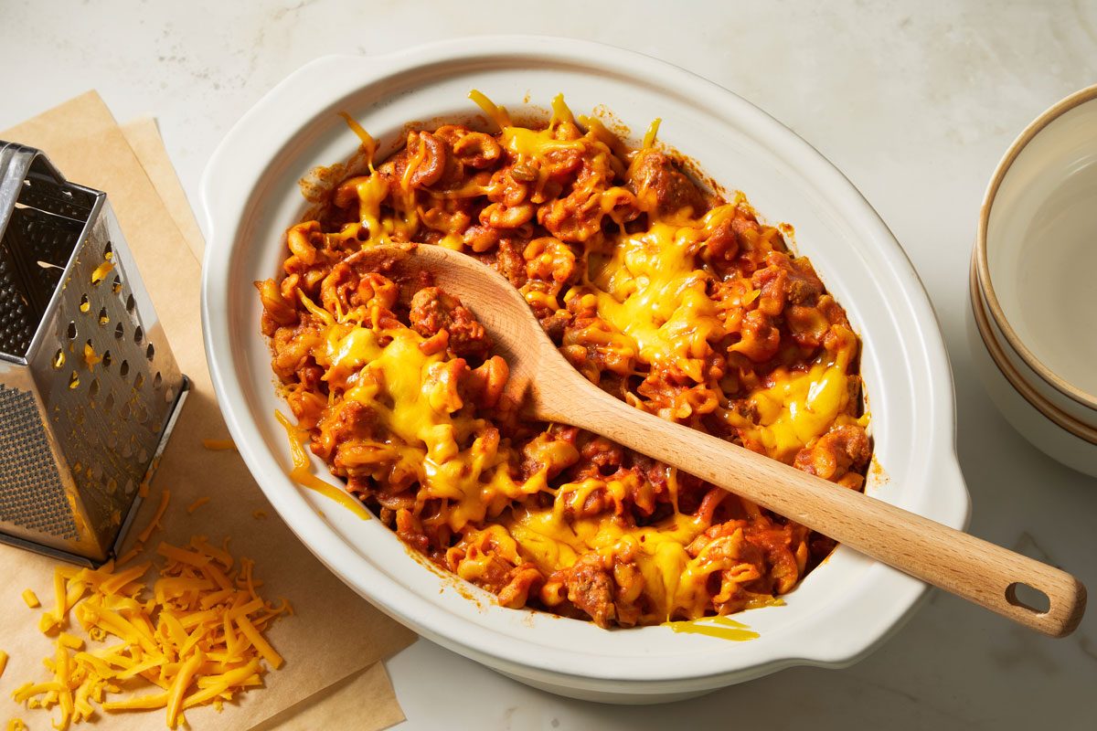 Overhead shot of Turkey Taco Macaroni; in a large slow cooker; top with cheese; empty bowls; serving spoon; marble surface