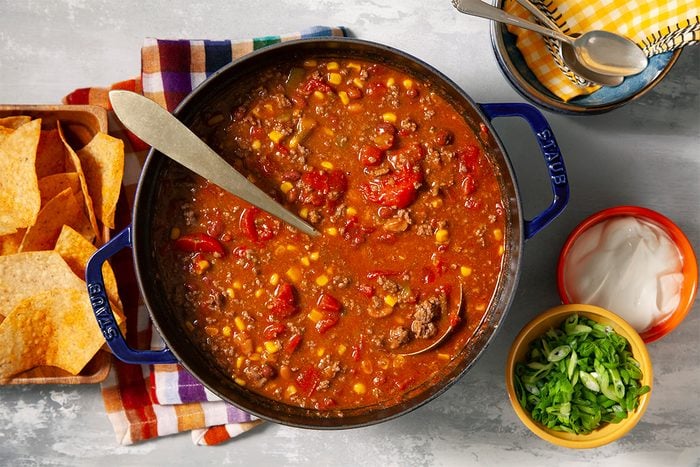 A pot of chili with tomatoes, corn, and beans, served with a ladle. Tortilla chips are on the side, and small bowls of chopped green onions and sour cream are ready for garnishing. A checkered napkin and spoons complete the setting.