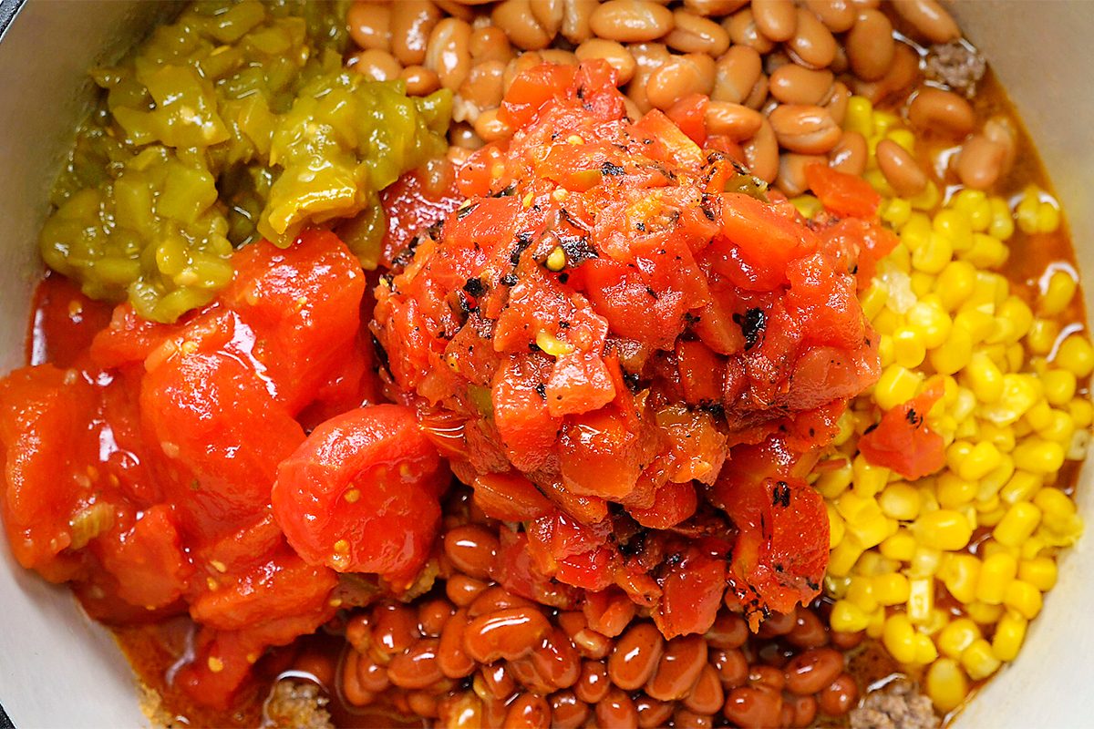 A colorful dish featuring chopped tomatoes, green chilies, pinto beans, corn, and more beans, likely a chili or stew, artfully mixed in a pot.