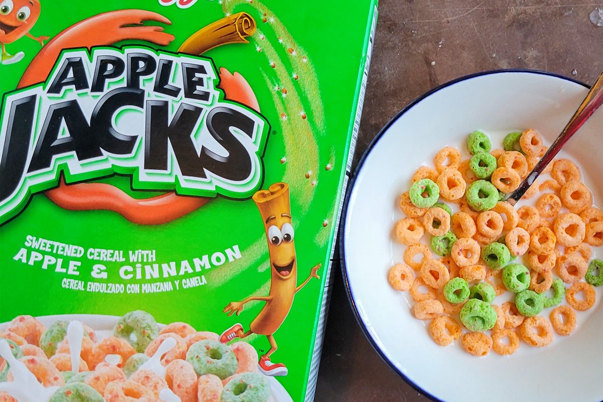 A box of Apple Jacks cereal is next to a bowl filled with the cereal and milk. The box features green and orange colors with animated apple and cinnamon stick characters. The cereal in the bowl contains green and orange loops.