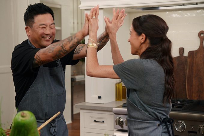 Meghan Markle and Roy Choi in matching aprons sharing a high-five and smiling in a kitchen. A large green vegetable is visible in the foreground. They appear to be celebrating or having fun while cooking.