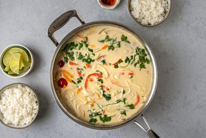 A pot of creamy soup with red and yellow bell peppers and garnished with fresh herbs. Accompanied by bowls of white rice and lime wedges on the side, set on a gray surface.