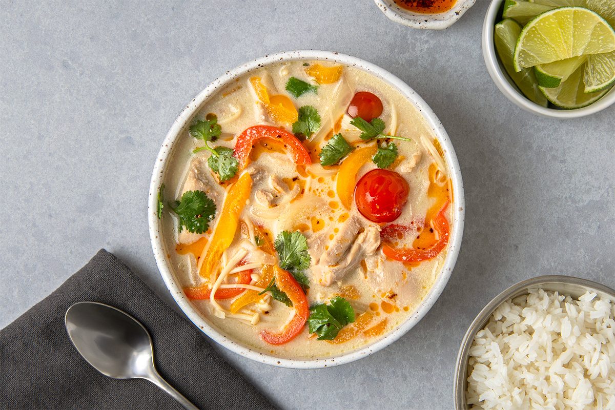 A bowl of creamy soup with chicken, red and yellow bell peppers, cherry tomatoes, and cilantro. Accompanied by a side of lime wedges, a small bowl of rice, and a metal spoon on a gray napkin.