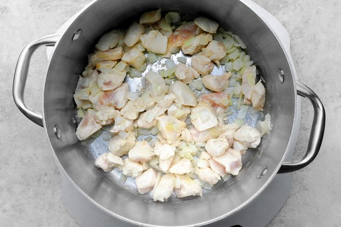 A steel pot on a stove containing chopped onions and diced chicken pieces being sautéed. The ingredients are lightly browned, creating a savory base. The pot has sturdy handles on each side.