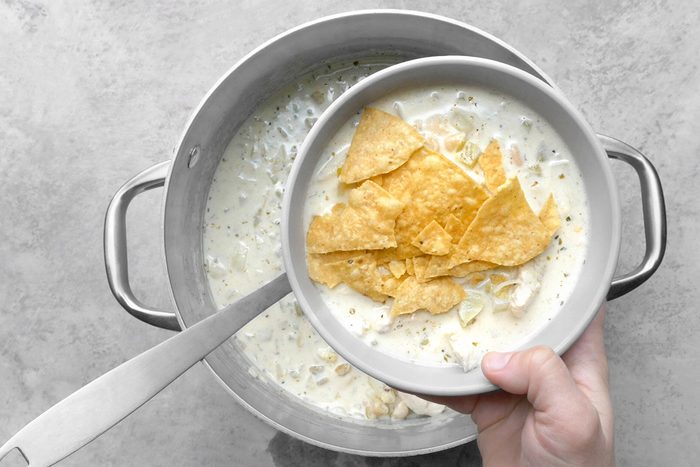 A hand holding a bowl of creamy soup topped with broken tortilla chips. The bowl is being held over a pot filled with more of the same soup, and a ladle rests on the edge of the pot. The background is a light gray surface.