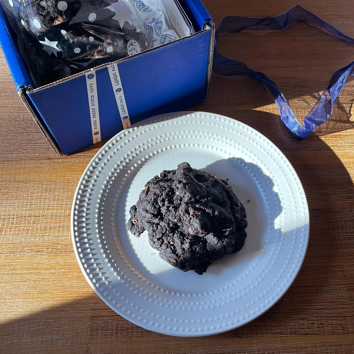 Dark brown cookie on a small white plate with a blue box and ribbon on a wooden table