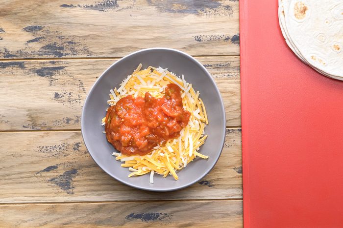 overhead shot of a bowl of shredded cheese and salsa sitting on a wooden table; next to the bowl on the right is a red cutting board with a stack of tortillas on it