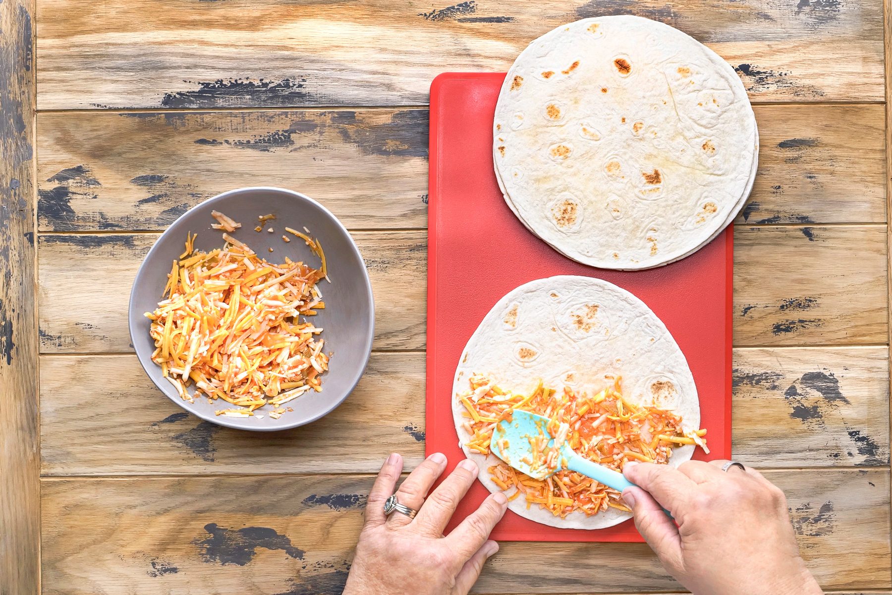 overhead shot of a person preparing a quesadilla; a flat, red cutting board sits on a wooden countertop and on it are two white tortillas; a second, gray bowl holds a mixture of shredded cheese and sauce; the person is spreading the cheesy filling onto one of the tortillas with a blue spatula