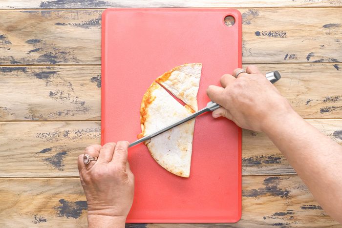 overhead shot of a person is cutting a quesadilla in half on a red cutting board; the quesadilla is on the board and the person is holding a silver knife to cut it; their left hand is holding the quesadilla in place;
