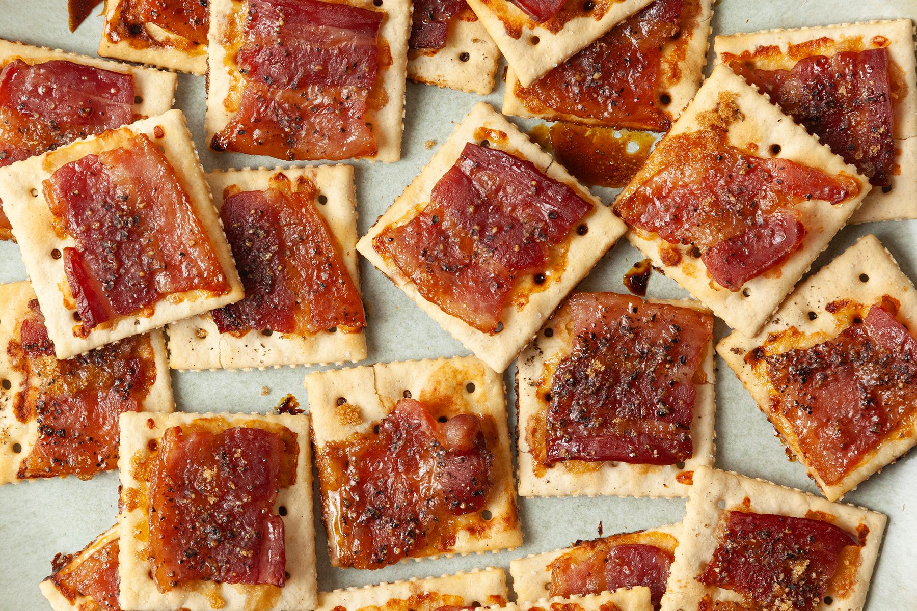 Square crackers topped with cooked bacon slices and a caramelized glaze are arranged in a scattered pattern on a light surface. The combination appears savory and crispy, with a golden-brown color on the bacon.