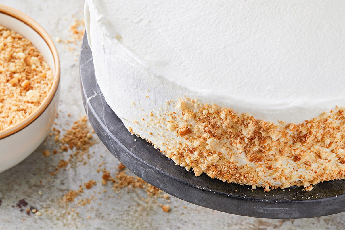 a closeup shot of a creamy a cake; it is presented on a round, dark stone plate; the cake has a smooth upper surface and is garnished with a golden crumb crust at its base; beside the cake is a bowl filled with more of the same crumb topping; the background shows a textured surface