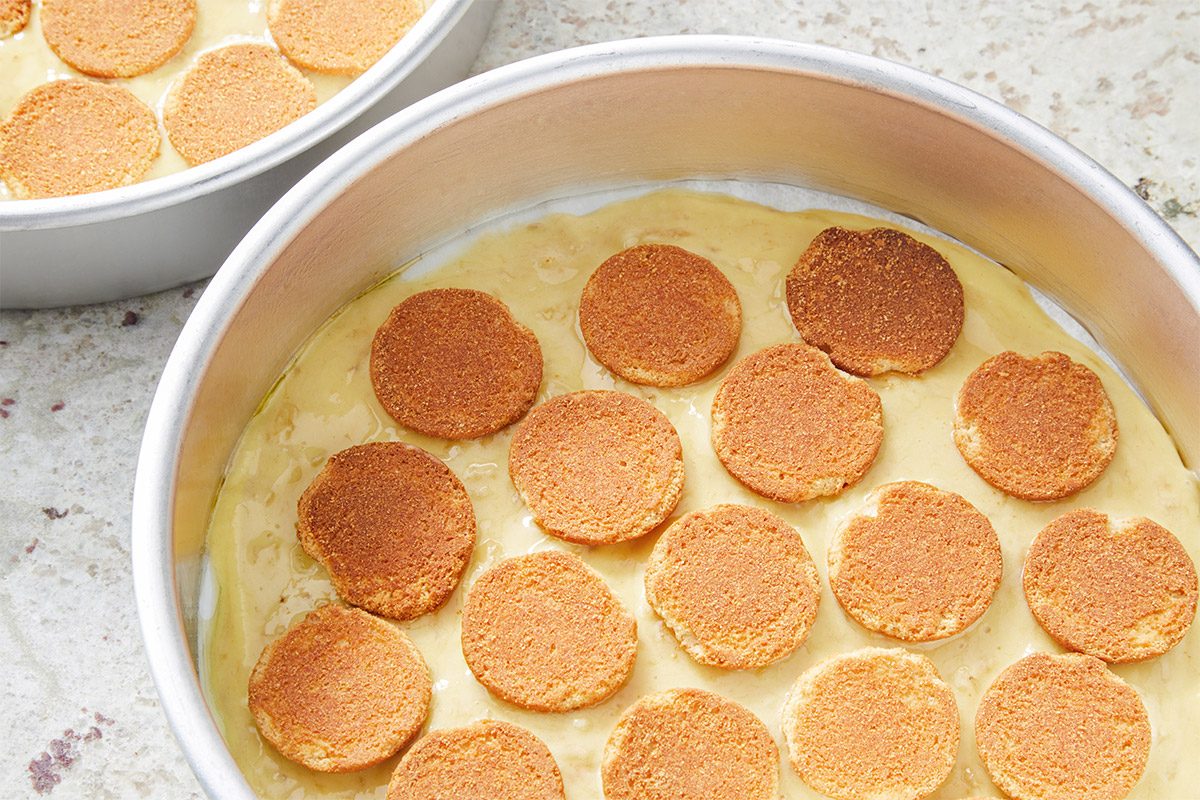 overhead shot of two round aluminum baking pans filled with a creamy, pale yellow mixture; round, golden-brown cookies are placed evenly on top of the batter in a pattern; the background features a light, textured surface