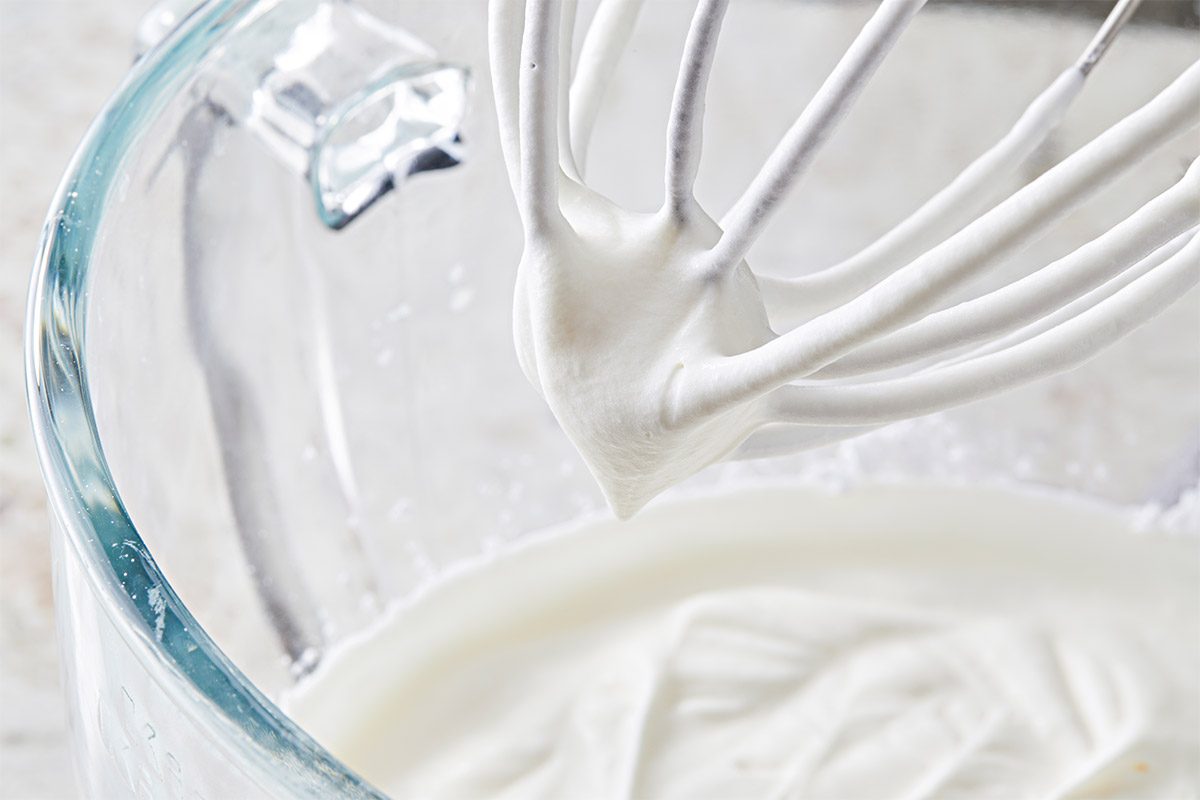 a closeup shot of a whisk attachment, showing the thick, fluffy mixture clinging to it, behind the whisk, a glass bowl partially filled with the same mixture is visible
