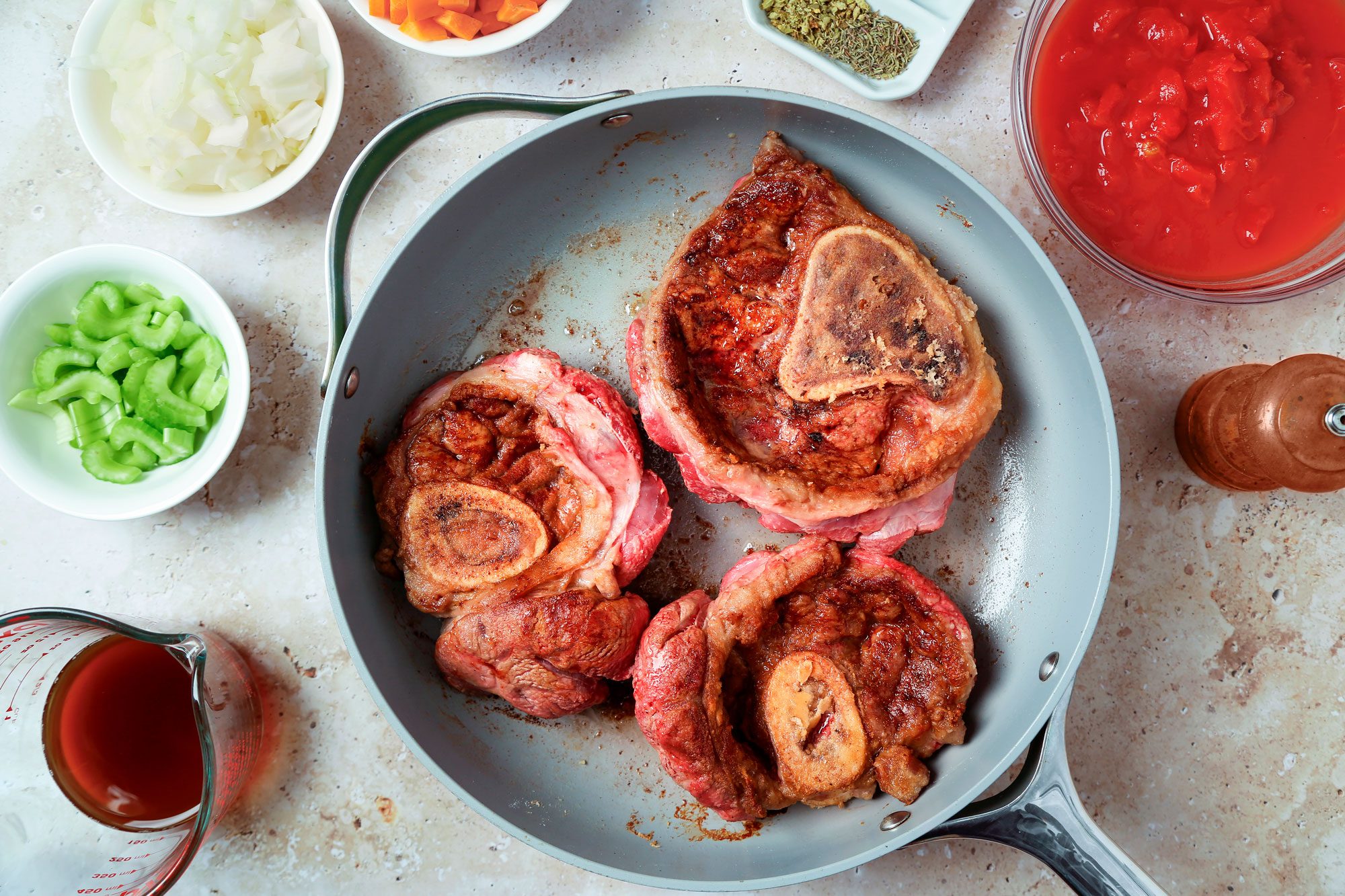 overhead shot of a kitchen counter with various ingredients and a pan containing osso buco, a pan of osso buco is sitting on the counter; there are also bowls of chopped onions, celery, and tomatoes; there is also a small bowl containing dried herbs, a small bowl containing diced carrots, a cup of red liquid, and a metal pepper grinder