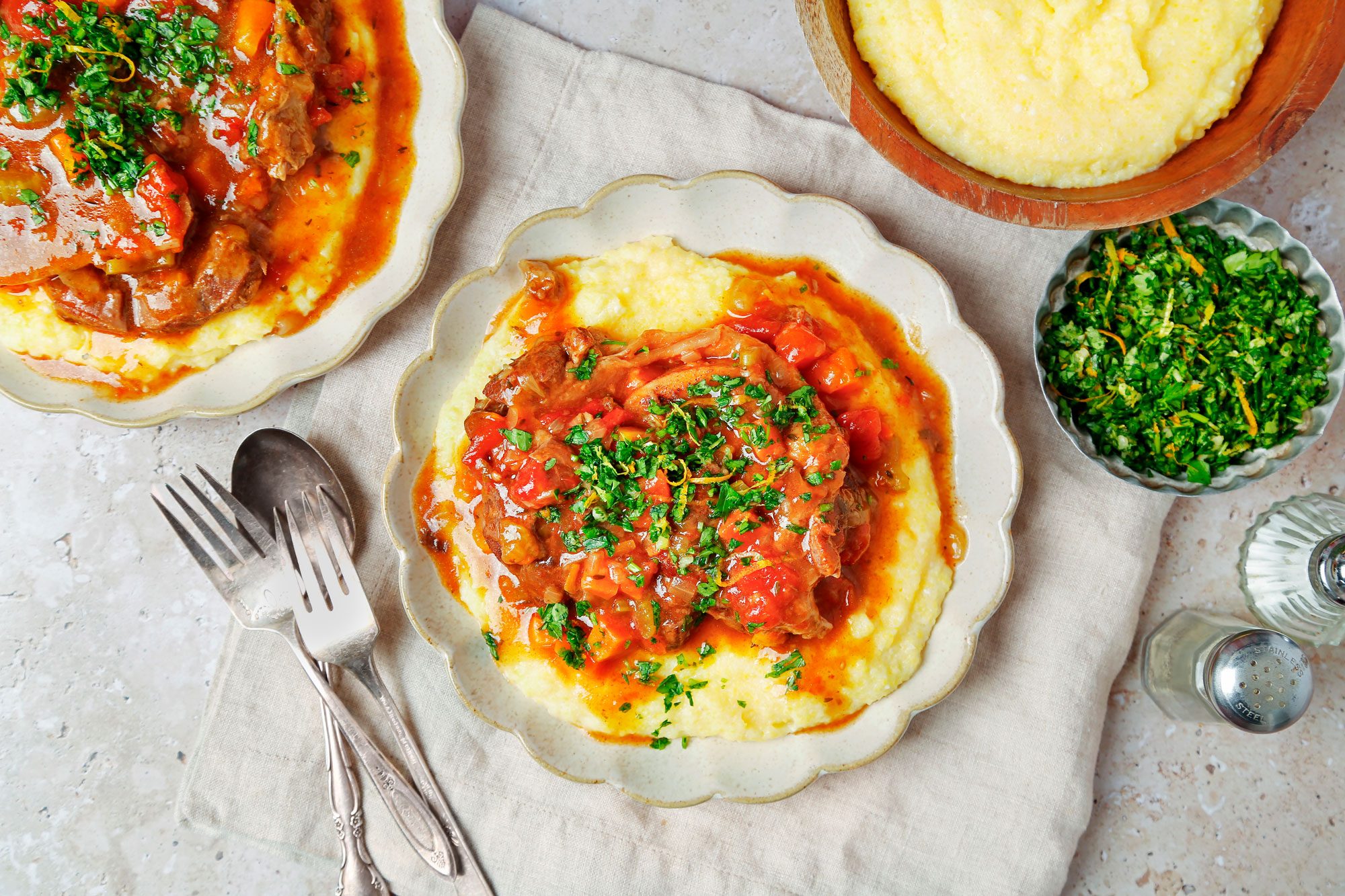 overhead shot of osso buco served over creamy polenta, garnished with fresh herbs and lemon zest; the meal is arranged on two plates and a bowl of polenta; two forks and a spoon are on a napkin beside the plates; there is also a bowl of chopped parsley and lemon zest and two salt shakers on the table; the background is a light brown with a beige tablecloth