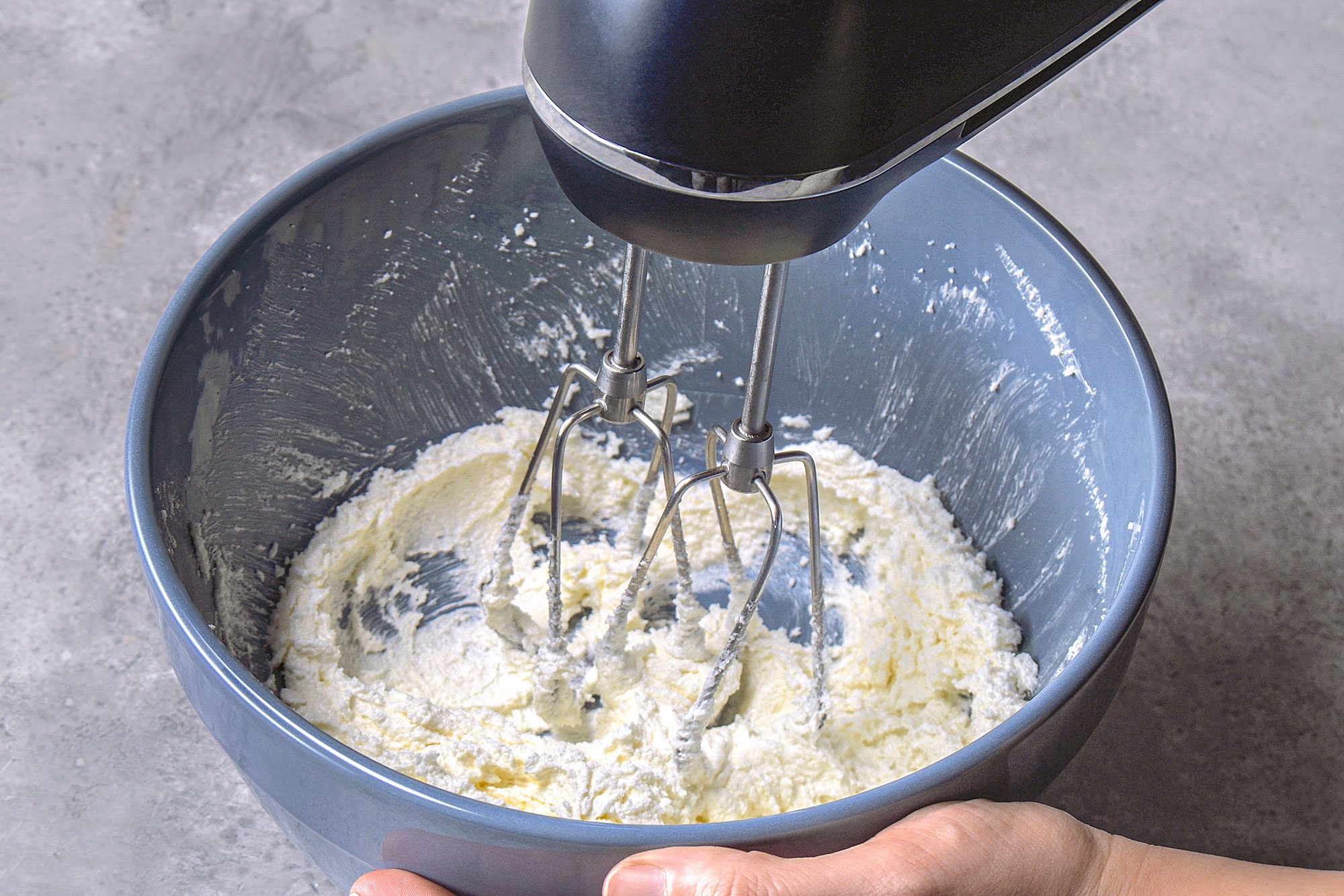 3/4th shot of a black hand mixer in operation inside a blue bowl, the bowl contains a mixture of creamy, white to yellowish ingredients, which are being whipped or blended by the mixer's beaters, the bowl is a light blue color sits on a textured grey surface