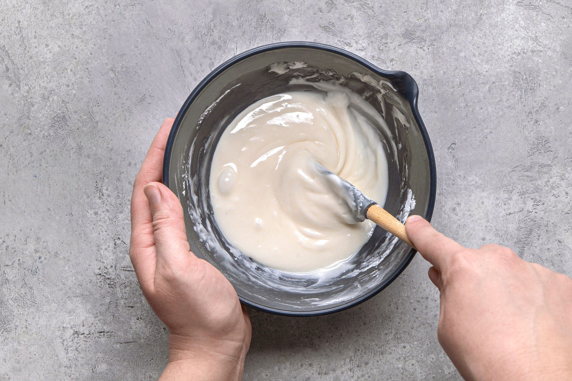 overhead shot of a pair of hands holding a blue bowl, inside which a smooth, white glaze is being stirred with a spatula, and the background is a textured grey surface