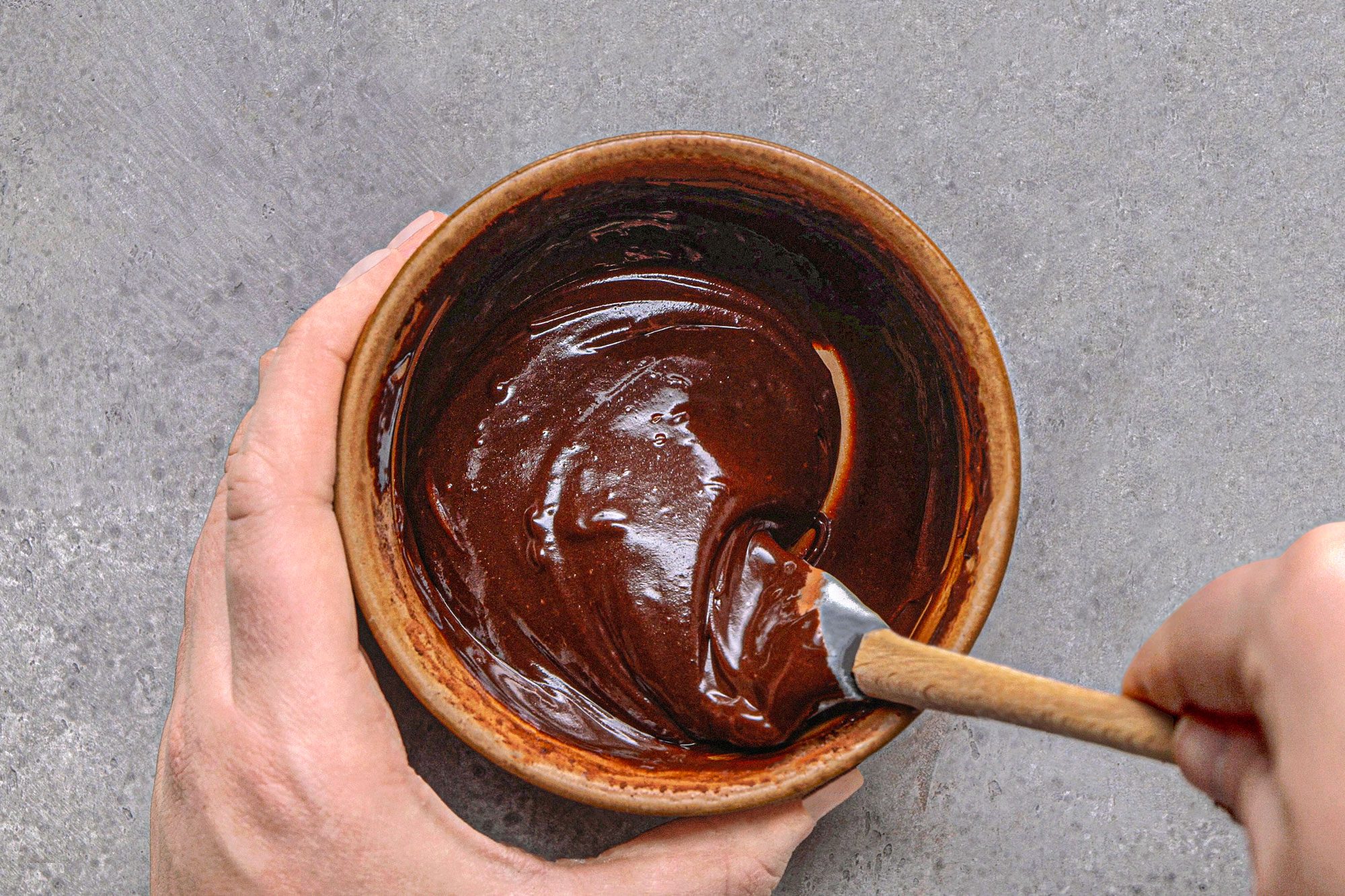 overhead shot of a pair of hands holding a brown bowl, inside which a chocolate ganache, is being stirred with a spatula, and sits on a textured grey surface