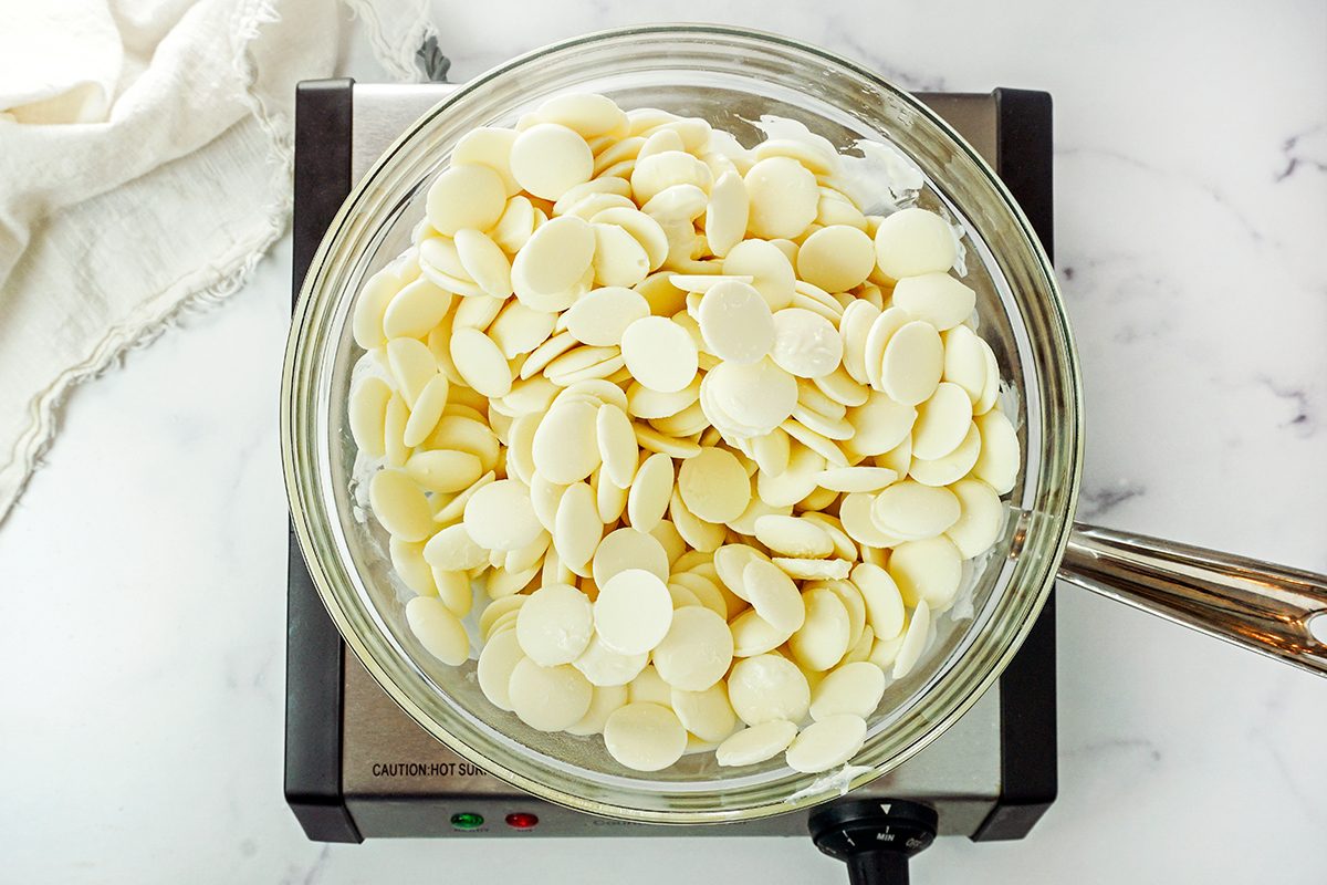 A glass bowl filled with white chocolate chips sits on an electric stovetop. A whisk is visible on the side, and a white cloth is partially seen in the background on a marble surface.
